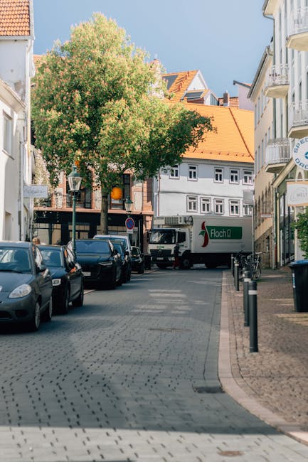 A male mover wearing a blue jacket and dark trousers is seen loading a large, light-colored sofa into a white delivery van parked on a city street. The back doors of the van are open, revealing a spac