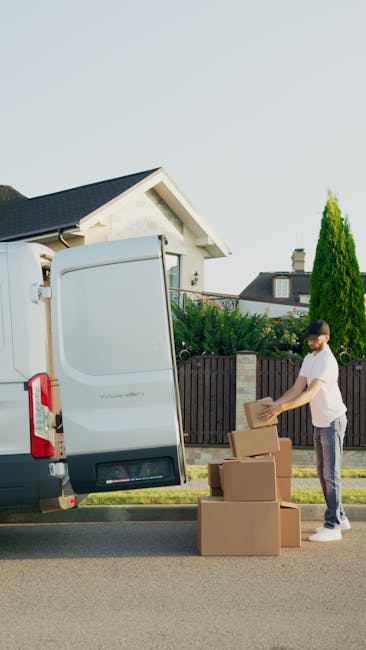 Two movers from Storage Hornchurch are loading a large, green upholstered furniture piece into the back of a white van parked on a paved area near a sidewalk. One worker, wearing a dark uniform, is li