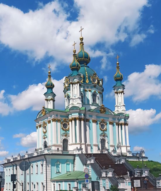 A stone church with a tall, crenellated tower is situated on a grassy area surrounded by gravestones of various shapes and sizes, some with moss or lichen growth. The church is built from grey stone w