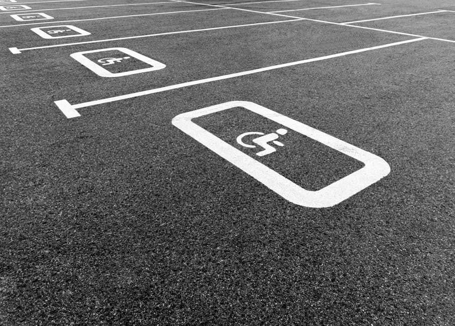 An asphalt parking area with clearly marked white lines and designated spaces, including a prominent white symbol indicating accessible parking for disabled individuals, with additional parking bays v