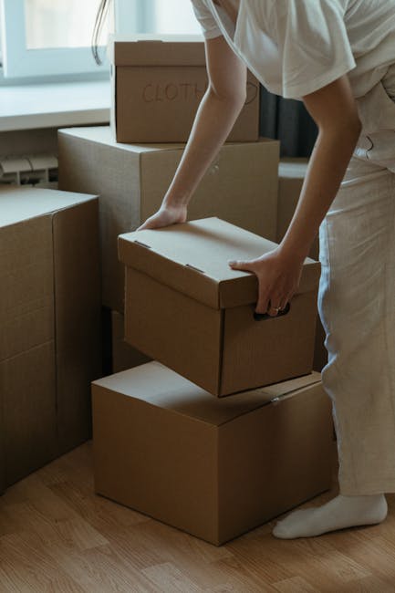 A woman sitting cross-legged on a wooden floor inside a room surrounded by stacked cardboard moving boxes of various sizes, some sealed with red or black tape. She is holding and tossing packing polys